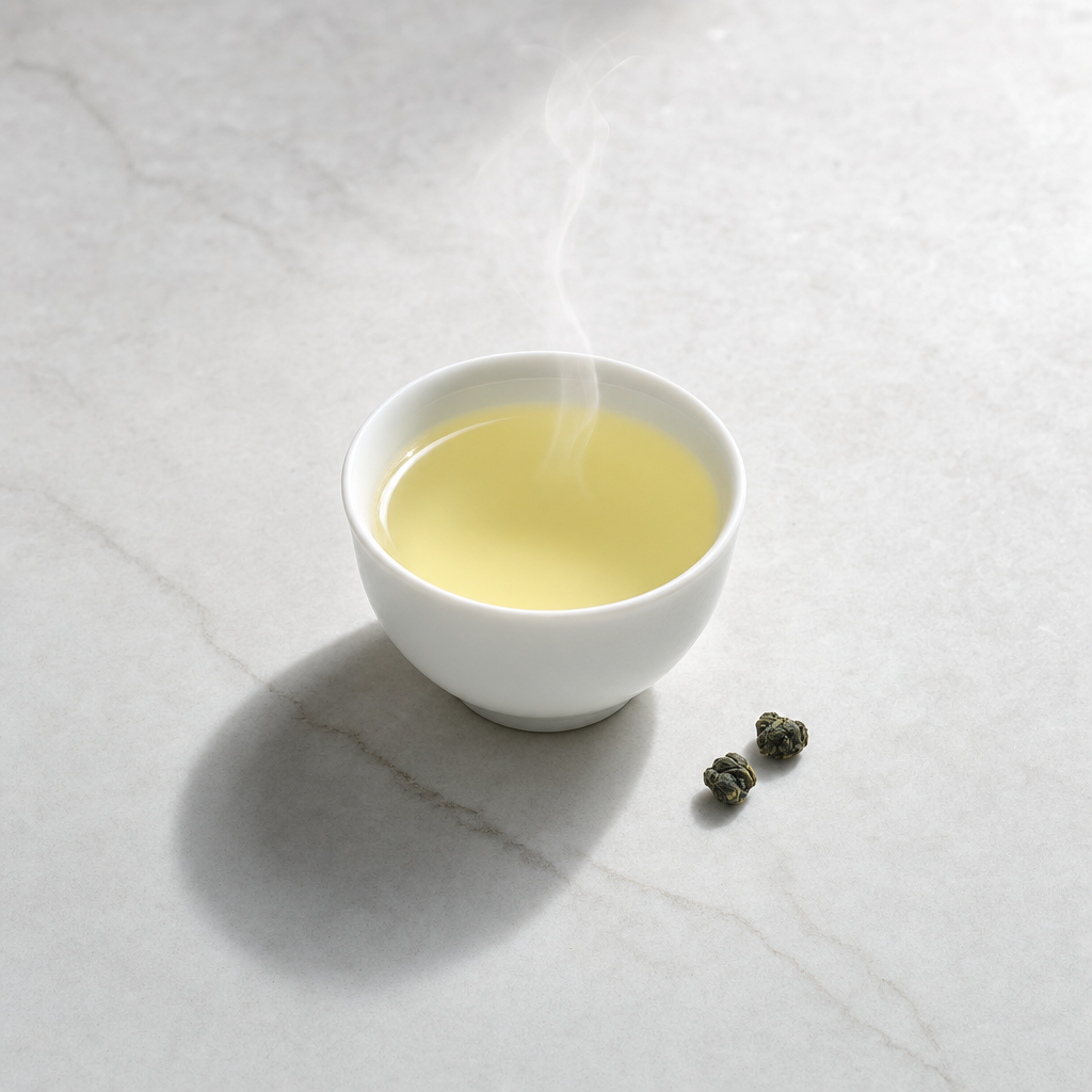 A small white porcelain cup of pale jade-yellow Mountain Crown Reserve Oolong liquor on a whitish-grey marble surface, with two rolled pellets beside it