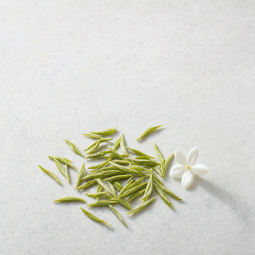 A small relaxed scatter of loose slim green Huatan Signature Jasmine tea leaves on a pale grey stone surface beside a white jasmine blossom