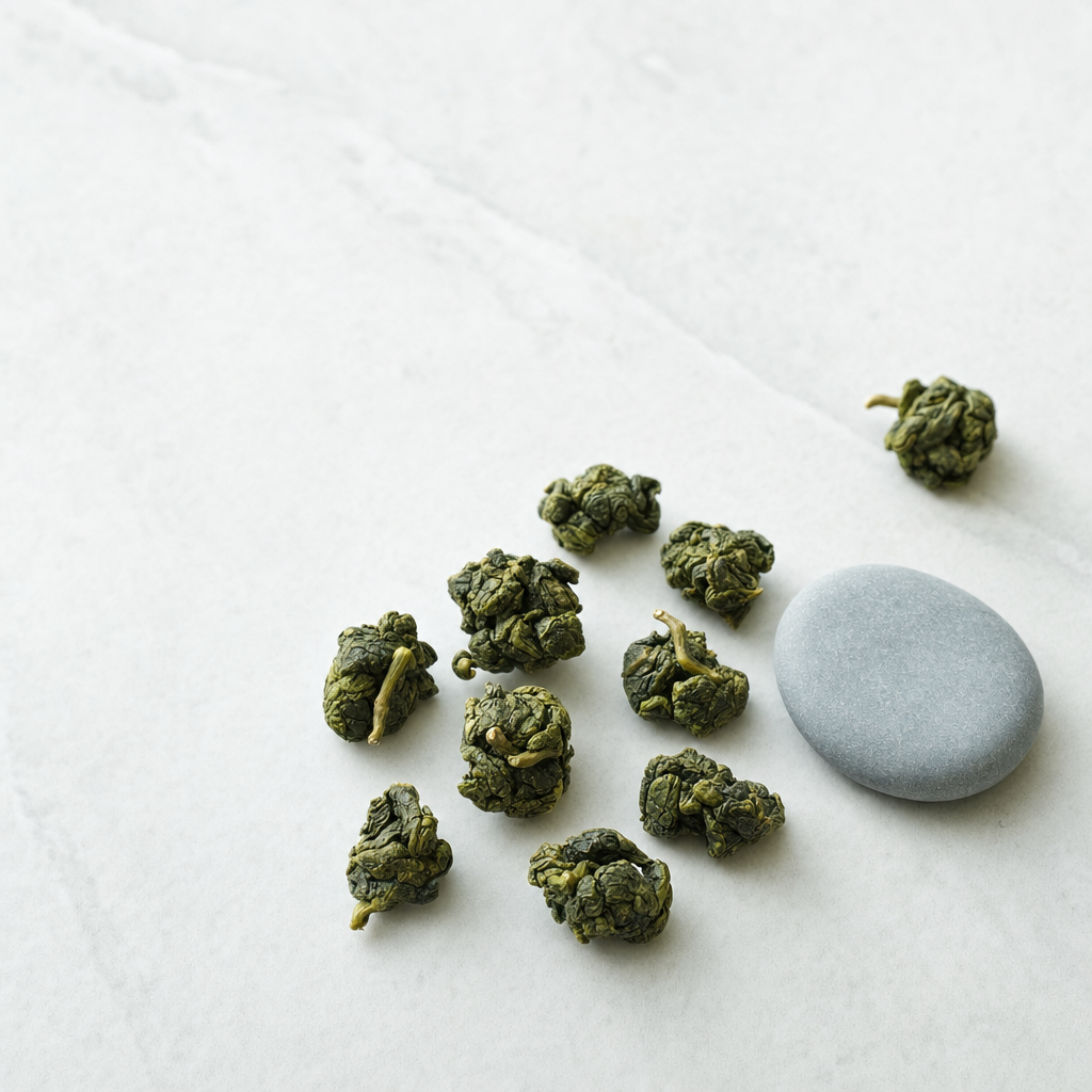 A small relaxed scatter of rolled Mountain Crown Signature Oolong pellets on a pale grey stone surface beside a smooth blue-grey pebble