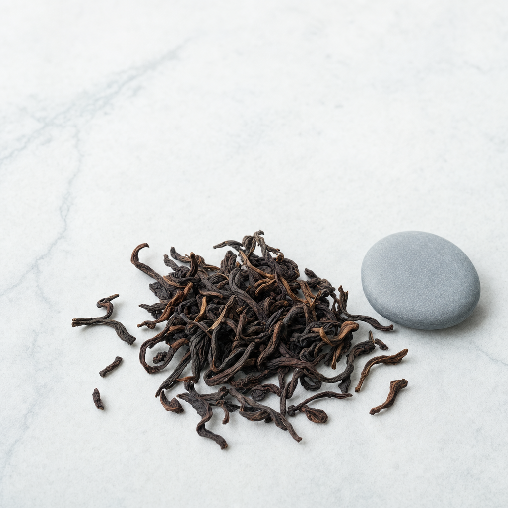 A small relaxed scatter of loose dark reddish-brown Yunnan Crown Signature Pu-erh leaf on a pale grey stone surface beside a smooth blue-grey pebble