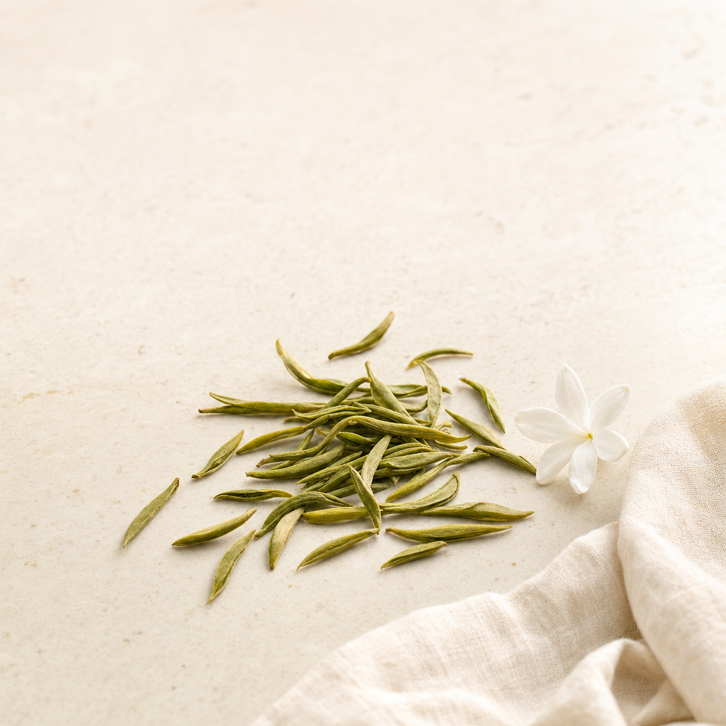 A small arrangement of loose slim green Huatan Select Jasmine tea leaves on a warm cream stone surface beside a white jasmine blossom and a soft fold of linen
