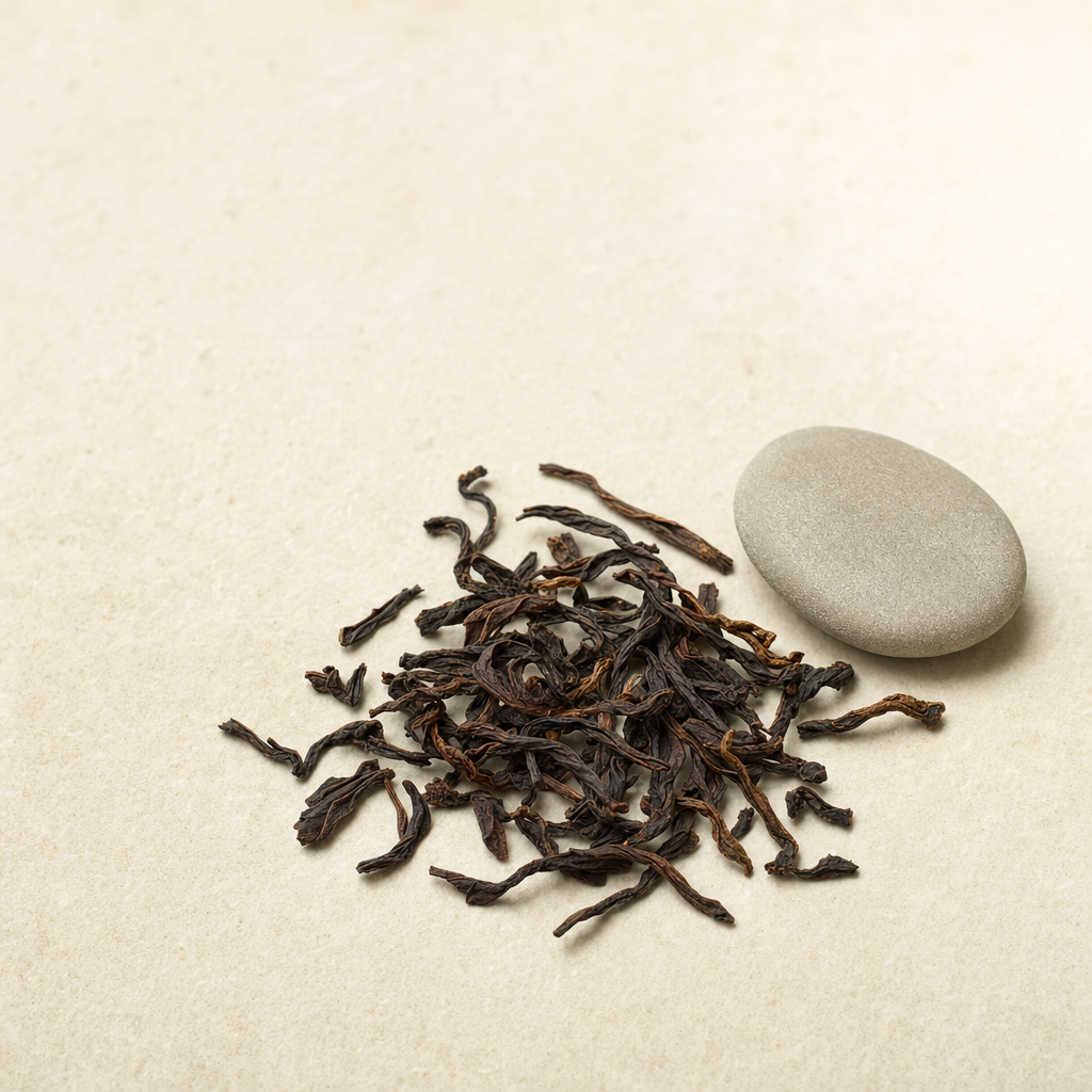 A small relaxed scatter of loose dark reddish-brown Yunnan Crown Reserve Pu-erh leaf on a pale stone surface beside a smooth warm-grey pebble