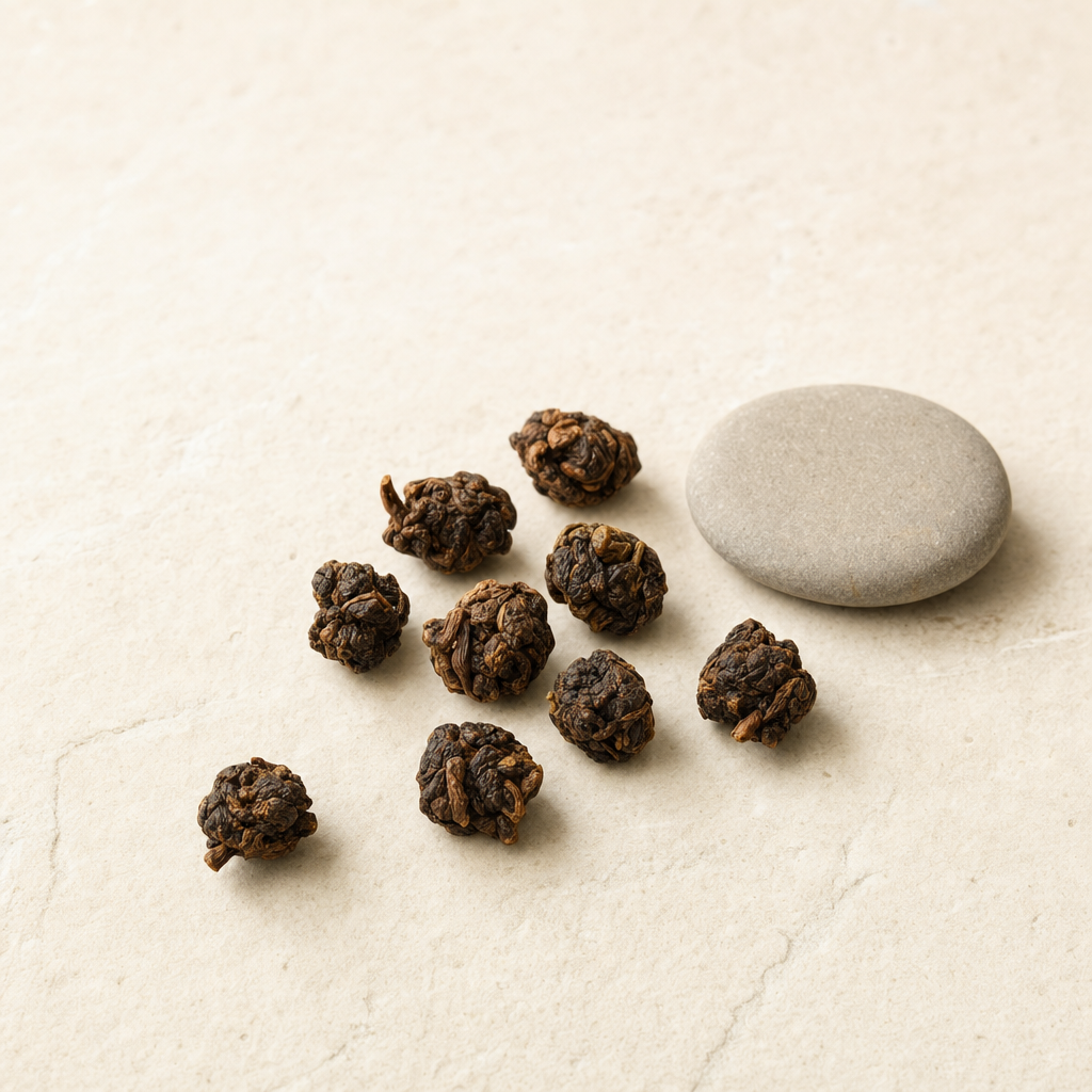 A small relaxed scatter of roasted amber-brown Ember Crown Reserve Dark Oolong pellets on a pale stone surface beside a smooth warm-grey pebble