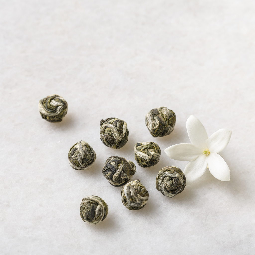 A small relaxed scatter of hand-rolled silvery-green Pearl Jasmine tea pearls on a pale stone surface beside a white jasmine blossom