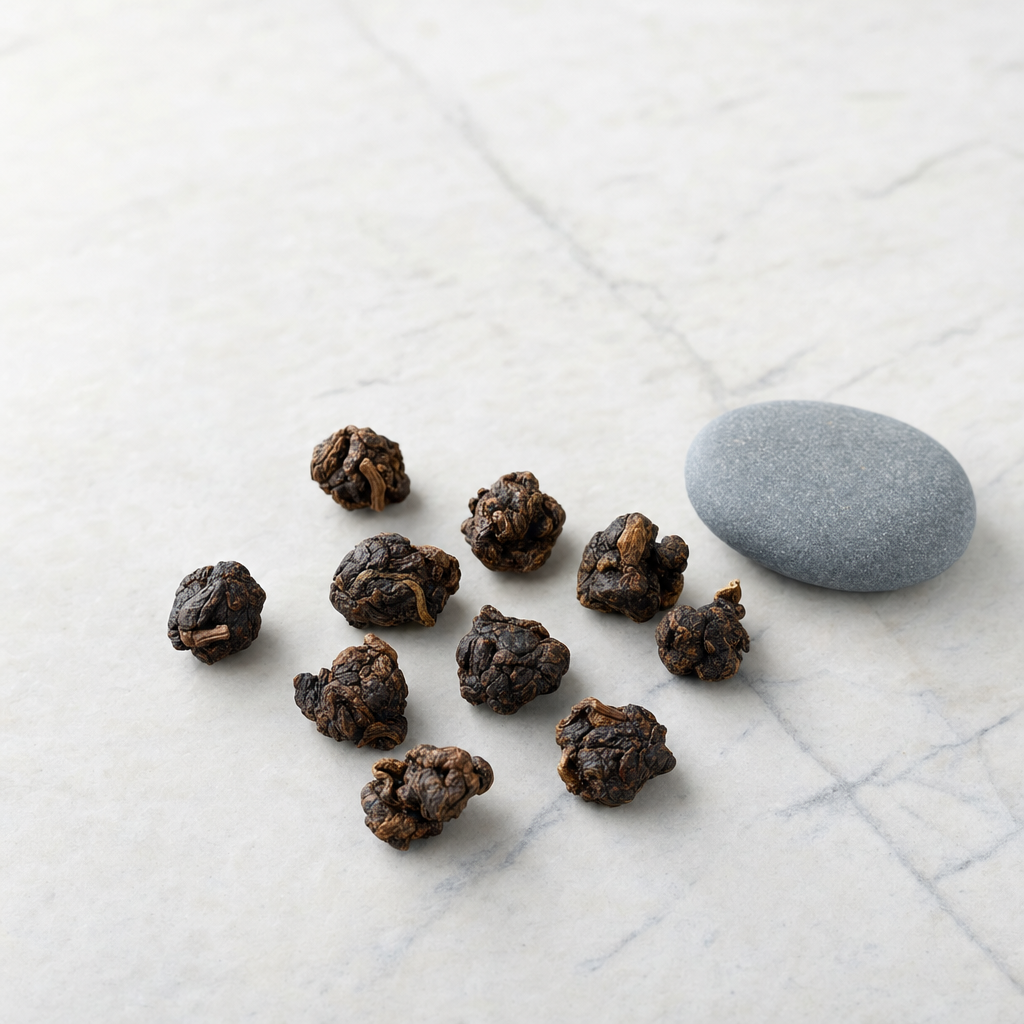 A small relaxed scatter of roasted amber-brown Ember Crown Signature Dark Oolong pellets on a pale grey stone surface beside a smooth blue-grey pebble
