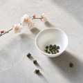 Mountain Crown Reserve Oolong loose-leaf rolled jade pellets in a white porcelain bowl on a bright stone surface, with a plum blossom branch