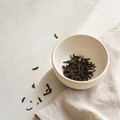 Yunnan Crown Select Pu-erh loose dark reddish-brown leaf in a small cream stoneware cup on a bright stone surface, with a soft folded linen