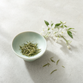 Huatan Signature Jasmine loose slim green tea leaves in a pale celadon bowl on a bright stone surface, with fresh white jasmine blossoms