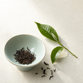 Yunnan Crown Signature Pu-erh loose dark reddish-brown leaf in a pale celadon bowl on a bright stone surface, with a fresh tea sprig