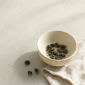 Mountain Crown Select Oolong loose-leaf rolled jade pellets in a small cream stoneware cup on a bright stone surface, with a soft folded linen