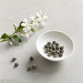 Pearl Jasmine hand-rolled green tea pearls in a small white porcelain bowl on a bright stone surface, with fresh white jasmine blossoms
