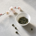 Mountain Crown Reserve Oolong hand-rolled jade pellets in a small white porcelain bowl on a pale stone surface, with a fresh plum blossom branch curving into the frame